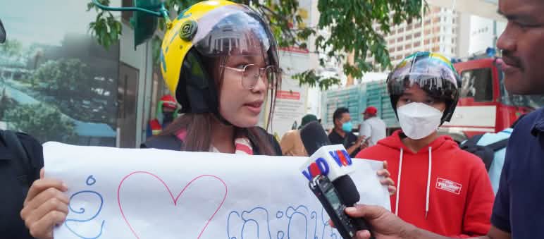 „Wij houden van Koh Kong Island!”: Campagne van Mother Nature Cambodia ter bescherming van het eiland tegen uitbuiting Een jonge vrouw met kleurrijke helm houdt een poster met hart omhoog en spreekt in een microfoon
