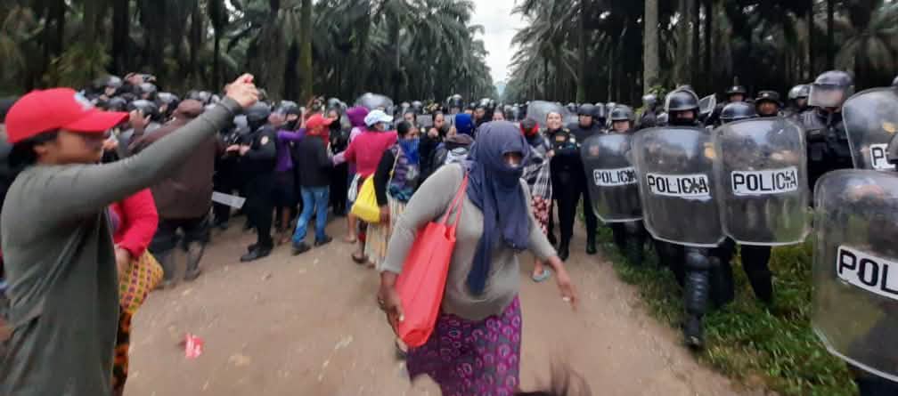 Een groep Maya's protesteert aan de linkerkant van de foto, terwijl politie-eenheden met helmen en schilden oprukken onder oliepalmen aan de rechterkant van de foto