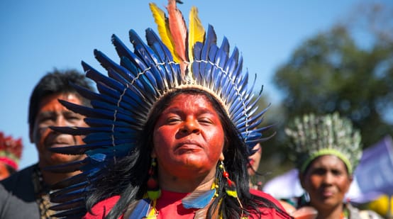 Een met een verenkroon versierde inheemse vrouw; op de achtergrond twee andere vrouwen
