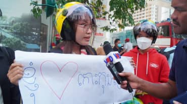 Een jonge vrouw met kleurrijke helm houdt een poster met hart omhoog en spreekt in een microfoon