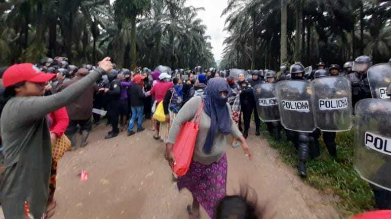 Een groep Maya's protesteert aan de linkerkant van de foto, terwijl politie-eenheden met helmen en schilden oprukken onder oliepalmen aan de rechterkant van de foto