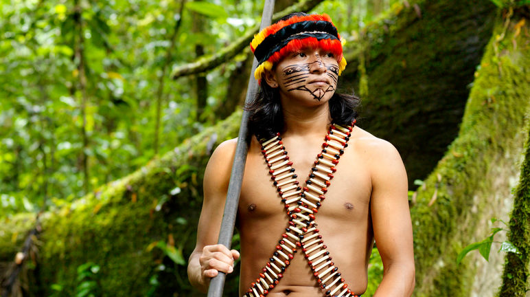 Een inheemse man die versierd is met veren en beschilderingen in zijn gezicht heeft staat met een blaaspijp in het regenwoud